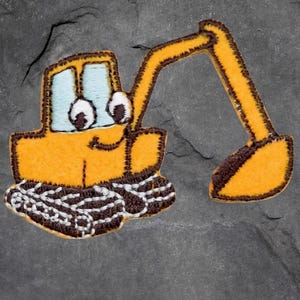May include: An embroidered patch featuring a cartoon excavator in yellow and brown. The excavator has a smiling face, blue window, and large white eyes. It has a long arm with a scoop. The patch is set against a grey background.
