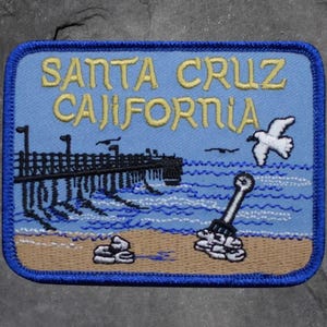 May include: Embroidered patch with a blue border and a light blue background. The patch features the text "SANTA CRUZ CALIFORNIA" in yellow, along with a pier, waves, a seagull, and a beach scene. The patch is rectangular with rounded corners.