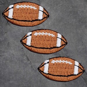 May include: Three embroidered football patches. Each patch features a brown football with white stripes and white stitching. The patches are outlined in black and are arranged diagonally on a grey surface.
