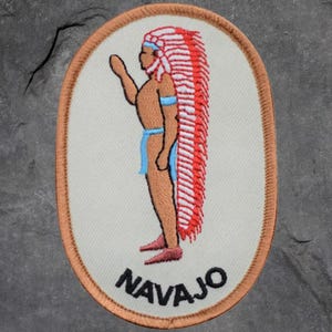 May include: Embroidered patch featuring a Native American figure in profile, wearing a red and white headdress and blue accents. The word "NAVAJO" is embroidered in black at the bottom, set against an off-white background with a tan border.