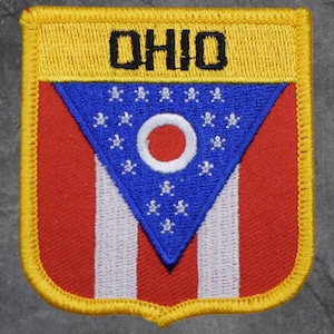 May include: Embroidered patch featuring the Ohio state flag design. The patch has a yellow border and a rectangular top section with the word "OHIO" in black. The main design includes a blue triangle with a white circle and stars, set against a red and white striped background.