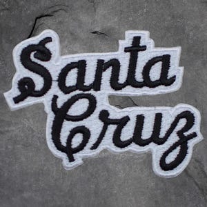 May include: Embroidered patch with the words "Santa Cruz" in a stylized script. The text is black with a white border and a light gray background. The patch has a textured appearance.