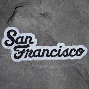 May include: A black and white embroidered patch with the words "San Francisco" in a cursive font. The text is black with a white border. The patch is on a gray, textured surface.