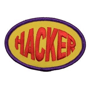 May include: Embroidered patch featuring the word "HACKER" in red, bold lettering. The text is set on a yellow oval, with a purple border. This patch is designed for application on clothing or accessories.