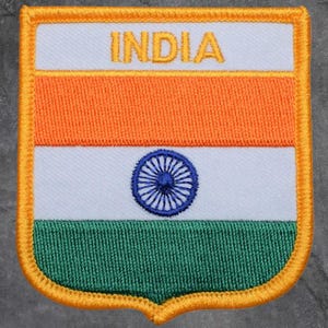 May include: Embroidered patch depicting the Indian flag. The patch has a white background with an orange stripe at the top, a blue wheel in the centre, and a green stripe at the bottom. The word "INDIA" is embroidered in gold.