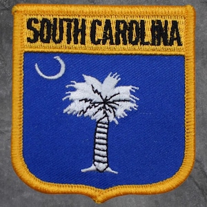 May include: A shield-shaped patch featuring the words "SOUTH CAROLINA" in black on a gold border. The patch has a blue background with a white crescent moon and a white palmetto tree, a symbol of the state.