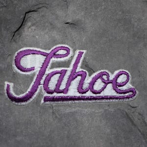 Könnte beinhalten: Gestickter Aufnäher mit dem Wort "Tahoe" in Schreibschrift. Die Buchstaben sind lila mit weißer Füllung und einem lila Rand. Der Aufnäher befindet sich auf einer grau strukturierten Oberfläche.