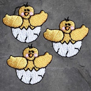 May include: Three yellow embroidered chicks hatching from white eggs. The chicks have black eyes, pink cheeks, and open beaks. The eggs are cracked open, revealing the chicks inside.