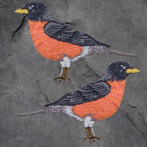 May include: Two embroidered robin birds with black, grey, white and orange details on a grey slate background.
