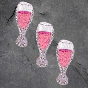 May include: Three embroidered patches shaped like wine glasses. Each patch features a pink liquid, a white rim, and a detailed stem. The patches are set against a dark gray, textured background.