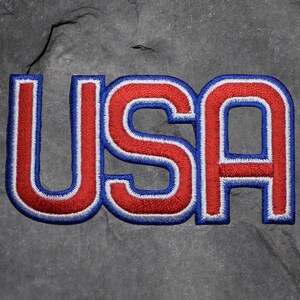 May include: Embroidered patch featuring the word "USA" in bold, capital letters. The letters are red with a white border and a blue outline. The patch is likely for clothing or accessories.