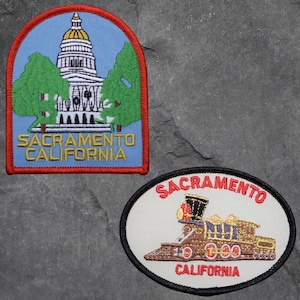 May include: Two embroidered patches with the text "Sacramento California". The first patch features a blue background with a white building and green trees. The second patch features a white background with a brown train and the text "Sacramento California".