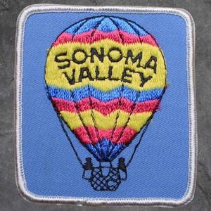 Könnte beinhalten: Gestickter Aufnäher mit einem bunten Heißluftballon-Design. Der Ballon ist gelb, rosa und blau, mit dem Schriftzug "SONOMA VALLEY" in Gelb. Der Aufnäher hat einen blauen Hintergrund und einen weißen Rand.