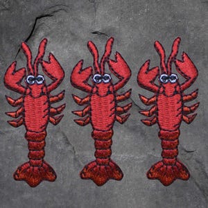 May include: Three embroidered red lobster patches with googly eyes. The patches feature a detailed design of the lobster's body, claws, and tail. The patches are likely for clothing or accessories.