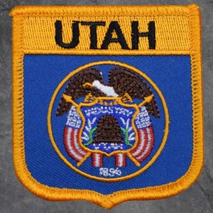 May include: Embroidered patch featuring the word "UTAH" in black on a gold background. The patch has a blue center with the state seal, including an eagle, beehive, and the year 1896. The patch is bordered with gold stitching.