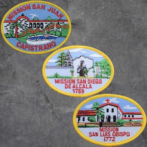 May include: Three embroidered patches depicting California Missions. The top patch is yellow with a blue background and reads "MISSION SAN JUAN CAPISTRANO". The middle patch is yellow with a blue background and reads "MISSION SAN DIEGO DE ALCALA 1769". The bottom patch is yellow with a blue background and reads "MISSION SAN LUIS OBISPO 1772".