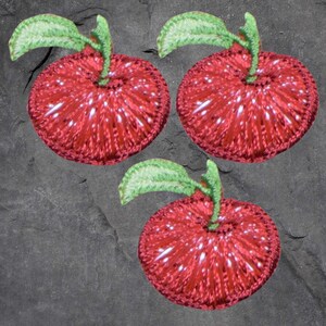 May include: Three crocheted red apple ornaments with green leaves and stems. The apples have white accents and are set against a dark gray background. These handmade ornaments are perfect for adding a touch of whimsy to any decor.