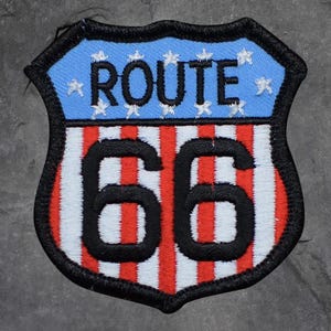 Könnte beinhalten: Gestickter Route 66-Aufnäher in Schildform. Der obere Teil ist hellblau mit weißen Sternen und dem Wort "ROUTE" in Schwarz. Der untere Teil hat rote und weiße vertikale Streifen mit der Zahl "66" in Schwarz.