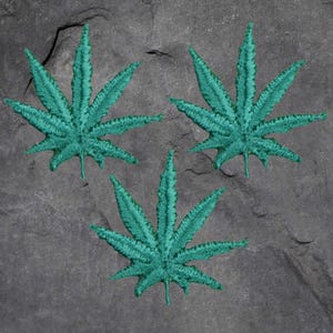 May include: Three embroidered teal cannabis leaf patches on a gray stone surface. Each leaf has detailed stitching, showcasing the veins and shape of the plant. The patches are evenly spaced, creating a visually appealing arrangement.