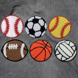 May include: Six different sports balls, each with a unique design and color. The balls are made of a soft, fuzzy material and are arranged in two rows of three. The balls include a baseball, soccer ball, softball, football, basketball, and volleyball.