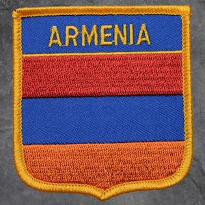 May include: Embroidered patch of the Armenian flag. The patch features a blue top section with the word "ARMENIA" in gold, a red middle section, and an orange bottom section. The patch is edged with gold stitching.