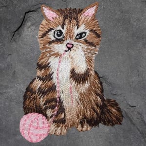 May include: Embroidered patch of a kitten playing with a ball of pink yarn. The cat has brown and white fur with pink accents on the ears and nose. The patch is designed to be sewn or ironed onto fabric.