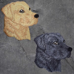 May include: Two embroidered dog portraits on a gray slate background. The dog on the left is a light brown Labrador Retriever with its head turned to the right. The dog on the right is a black Labrador Retriever with its head turned to the right.