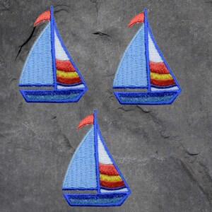 May include: Three embroidered sailboat patches. Each patch features a light blue sail with a white and striped red, yellow, and orange design. A small red flag is at the top of the mast. The boat's hull and outline are dark blue.