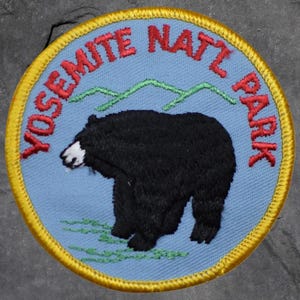 May include: Embroidered patch featuring a black bear with a white snout and green mountain range on a light blue background. The patch is circular with the words "YOSEMITE NATL PARK" in red, and a yellow border.