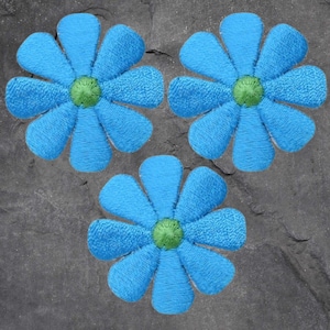 May include: Three embroidered blue flower patches with green centers are arranged on a gray stone surface. The flowers have a textured appearance, with each petal clearly defined. These patches could be used for clothing or craft projects.