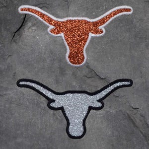 Könnte beinhalten: Zwei glitzernde Longhorn-Logos, eines orangefarben und eines silberfarben, werden vor einem dunkelgrauen, strukturierten Hintergrund angezeigt. Das orangefarbene Logo ist weiß umrandet, während das silberfarbene Logo schwarz umrandet ist. Beide Logos haben die gleiche Form.