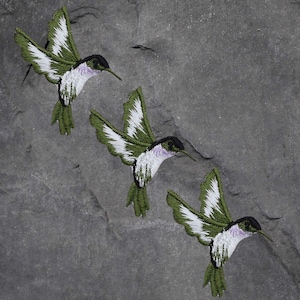 May include: Three green and white embroidered hummingbird patches on a grey background.
