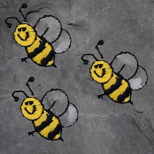 May include: Three embroidered bees with yellow and black striped bodies, smiling faces, and gray wings are displayed against a gray background. The bees are in a playful, cartoon style.