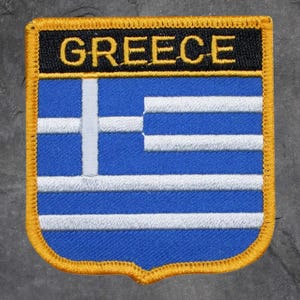 May include: Embroidered patch depicting the Greek flag. The patch features a blue field with white stripes and a white cross. The word "GREECE" is embroidered in gold lettering on a black banner at the top, with a gold border.