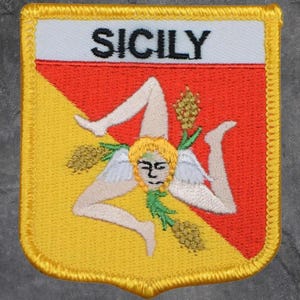 May include: Embroidered patch featuring the word "SICILY" above a triskeles symbol. The patch has a white top, red center, and yellow lower-left section. The triskeles has a central face with three legs extending outward, and wheat stalks.