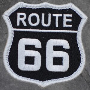 May include: A Route 66 patch featuring a black background with white embroidered text and border. The text "ROUTE" is at the top, and "66" is displayed below. The patch is shield-shaped.