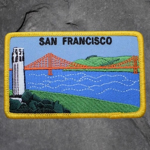 May include: Embroidered patch featuring the words "SAN FRANCISCO" above a depiction of the Golden Gate Bridge, a tower, and rolling green hills. The patch has a blue background with a yellow border.
