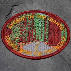 May include: Embroidered patch with an oval shape featuring a road winding through a forest of tall redwood trees. The patch has a red border and text that reads "AVENUE OF THE GIANTS, REDCREST, MYERS FLAT, PHILLIPSVILLE."