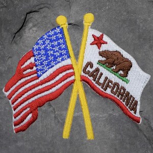 Könnte beinhalten: Gestickter Aufnäher mit der amerikanischen Flagge, die mit der kalifornischen Staatsflagge gekreuzt ist. Die amerikanische Flagge hat rote und weiße Streifen und ein blaues Sternenfeld. Die kalifornische Flagge zeigt einen Bären, einen Stern und das Wort "CALIFORNIA".