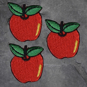 May include: Three embroidered apple patches. Each patch features a bright red apple with a yellow highlight and green leaves. The patches are arranged on a dark grey surface.