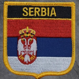 May include: Embroidered patch featuring the flag of Serbia. The patch has a black top with the word "SERBIA" in yellow. Below is a red stripe, a blue stripe, and a white stripe. The Serbian coat of arms is in the center.