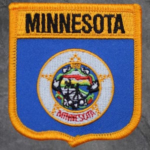 May include: A shield-shaped patch with the word "MINNESOTA" in black letters on a yellow background. The patch features a blue center with a circular emblem and the word "MINNESOTA" at the bottom.