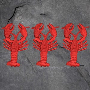Könnte beinhalten: Drei gestickte rote Hummer-Patches auf einem dunkelgrauen, strukturierten Hintergrund. Die Hummer sind detailliert mit Scheren, Beinen und Körpersegmenten dargestellt, was ein maritimes oder Küsten-Thema zeigt.