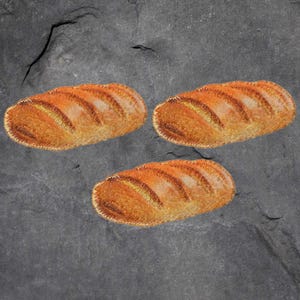 May include: Three embroidered bread loaves with golden-brown crusts and detailed stitching. The loaves are arranged on a dark grey, textured surface, creating a rustic and appetizing visual.
