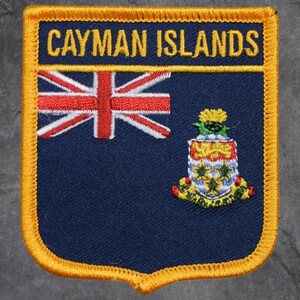 Peut inclure: Écusson bleu marine avec l'inscription "CAYMAN ISLANDS" en lettres dorées. L'écusson présente le drapeau Union Jack et les armoiries des îles Caïmans, avec une bordure dorée.