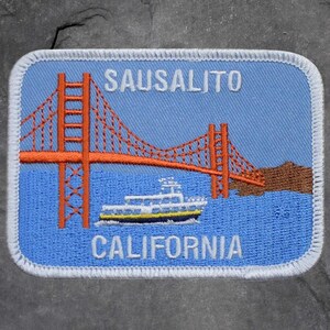 May include: Embroidered patch featuring the Golden Gate Bridge in orange, a white and yellow boat, and the text "Sausalito California" in white. The patch has a blue background and a white border.