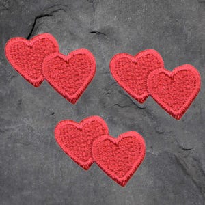 May include: Three sets of red heart-shaped embroidered patches. Each heart is a vibrant red with a textured appearance and a detailed border. The patches are arranged on a dark gray, textured surface, suitable for fabric embellishment.