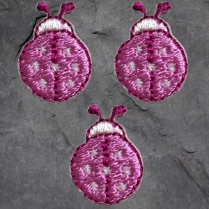 May include: Three pink embroidered ladybug patches with white accents. The ladybugs have white spots and are stitched with a pink thread.