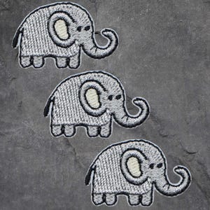 Könnte beinhalten: Drei graue gestickte Elefanten-Patches mit weißen Augen und schwarzen Umrissen. Die Elefanten sind vertikal in einer Reihe angeordnet.