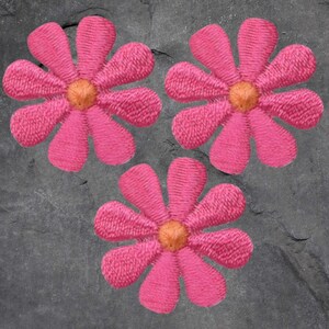 May include: Three embroidered pink daisy flower patches with orange centers. The flowers are arranged on a dark gray, textured surface. These patches are suitable for fabric crafts, adding a floral accent to clothing, bags, or other items.